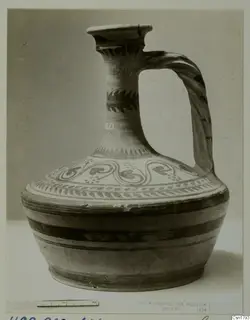 Painted jug with a tall neck, flared mouth and strap handle, shown against a grey backdrop