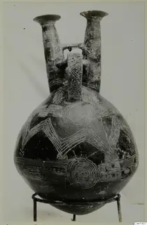 Double-spouted jug from Vounous with white incised zigzag and concentric circle patterns on a dark body.