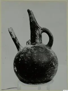 Red Polished III jug with a tall spout, a side spout and a loop handle, shown on a stand