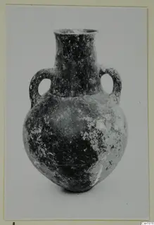 Two-handled pottery vessel with a tall neck and rounded body, from Vounous; dark surface with pale worn patches.