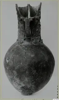 Globular vessel with a tall neck, two flaring openings and a bridge handle, dark worn surface, photographed on a plain background.