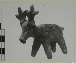 Clay figurine of an animal with branching antlers and four legs, photographed beside a scale bar.
