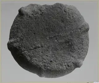 Stone fragment from Petra tou Limniti, roughly round with a rough surface, photographed against a plain grey background.
