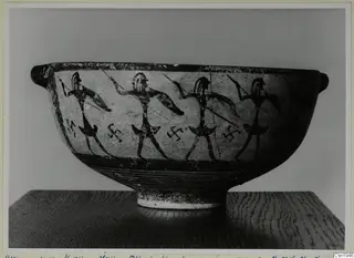 Black-on-Red I-III bowl with painted warriors carrying spears and shields, photographed on a wooden stand