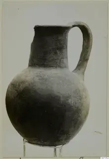 Dark, plain-surfaced pottery jug with a tall neck and strap handle, standing on a wire stand