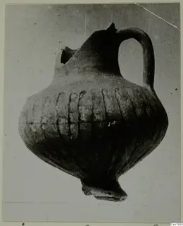 Pottery jug with a strap handle, grooved body and broken spout, shown against a grey backdrop