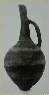 Dark pottery jug with a tall neck, flared lip and strap handle, shown against a grey background
