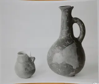 Two pottery jugs on a pale surface: a tall jug with a loop handle on the right and a small handled jug on the left.