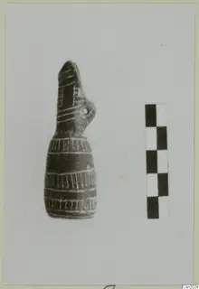 Dark pottery vessel with a tall beak-like top and pale banded lines, photographed beside a scale bar