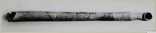 Long tubular object with a flared mouth at one end, dark grey surface with cracks, lying horizontally on a pale background