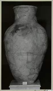 Large pottery pithos with a tall neck and incised ridged bands, standing on a plinth in a museum display.
