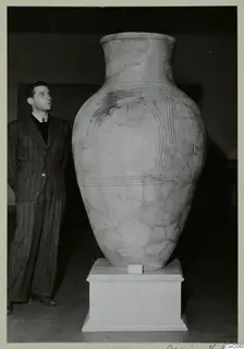 Large pale pottery vessel on a white pedestal, with a man in a striped suit standing beside it and looking up at it.