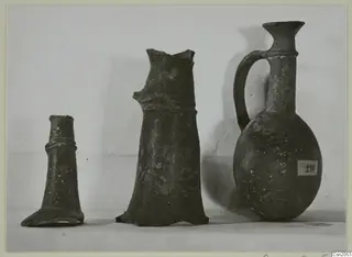 Three pottery vessels of Base-Ring Ware on a pale surface: two broken pieces and one complete juglet with a single handle.