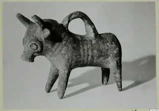 Pottery vessel modelled as a horned animal with a looped handle over its back, shown on a plain grey backdrop