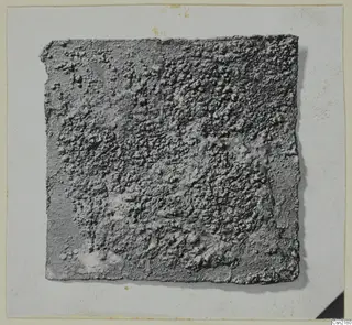 Corroded metal fragment, a flat square piece with a crusted grey surface, photographed on a card mount