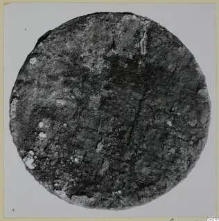 Circular corroded metal fragment with a faint grid-like pattern on its surface, photographed face-on