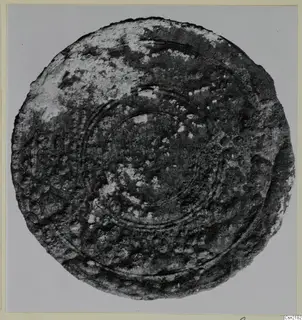 Corroded bronze disc with raised concentric rings and a pitted, bumpy surface, photographed on a grey backdrop.