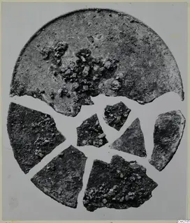 Corroded bronze lid with several broken fragments arranged around it, photographed on a pale grey background