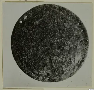 Round metal disc fragment with small rivet holes around its edge, photographed on a card mount