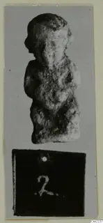 Small standing figure of a person with arms at the sides, photographed on grey card above a number label
