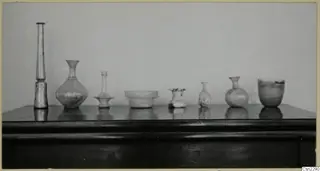 Seven blown glass vessels of varied forms standing in a row on a dark wooden table.