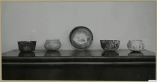 Five small vessels standing in a row on a dark shelf against a plain pale wall