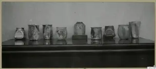 Row of weathered blown glass jugs and vases from Vouni, Korakas tombs, standing on a dark shelf.
