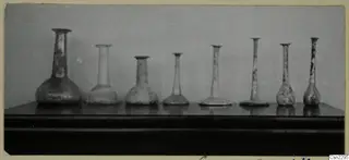 Row of several blown glass vessels with long slender necks standing on a dark shelf against a grey backdrop