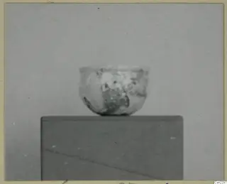Small thin-walled vessel with a wide mouth, standing on a dark block against a grey backdrop