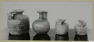 Four blown glass jugs and jars with handles standing in a row on a dark table.