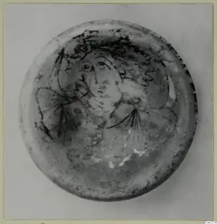 Round glass object with a dark painted face and hair pattern, photographed from above.