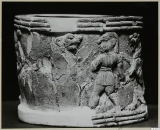 Cylindrical object with figures carved in relief on its side, showing a standing figure and an animal head