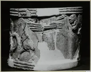 Cylindrical carved object with relief figures and ridged bands, photographed against a black background
