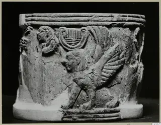 Carved cylindrical object with a relief of a winged creature and a seated figure, shown against a dark background
