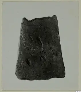 Corroded flat metal object with a broken top edge, photographed on a pale grey backdrop