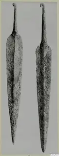 Two corroded metal daggers with long pointed blades and curled tangs, shown side by side.