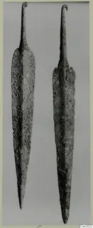 Two corroded metal weapons with long tapering blades and hooked tangs, shown side by side against a grey background.