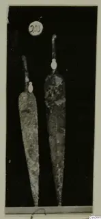 Two long pointed metal blades with narrow tangs, photographed upright against a black background.