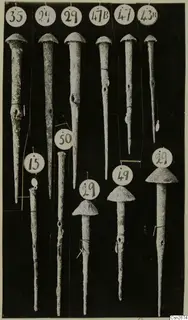 Eleven long metal implements with domed heads and tapering shafts, marked with round numbered paper labels, on a black background.
