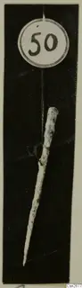 Slender metal tool with a pointed tip, hung by a thread from a round number label against a black background