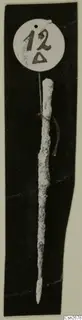Corroded bronze implement with a pointed tip, tied with string to a round number label, photographed on black.