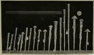 Row of about eighteen bronze tools with domed heads, standing upright on a wooden shelf against a dark background