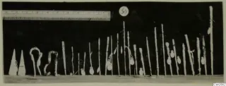 Row of bronze tools standing upright on a pale surface against a dark background, with a ruler and a number label