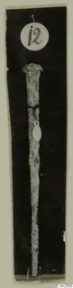 Slender bronze tool with a flattened head and tapering shaft, photographed upright with a round number label.