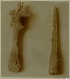 Two corroded bronze tools on a plain background: a larger one with a flared, curved upper end and a shorter, long pointed one beside it.