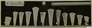 Twelve tapering bronze tools standing in a row against a dark background, with a ruler and a number card.