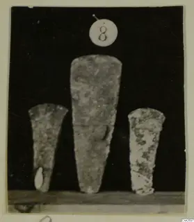 Three tapering tools standing upright on a shelf against a dark background, recorded as bronze tools