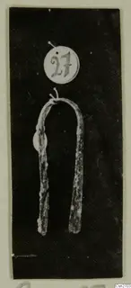 Bronze tool with two long arms joined by a curved bend, shown with a round number label, on a dark background.