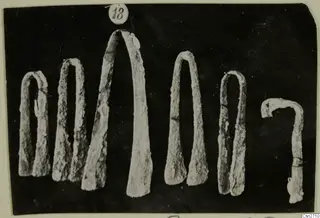 A row of corroded bronze tools with tapering arched arms against a dark background.