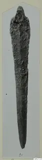 Long corroded bronze tool with a tapering blade and rounded upper end, shown on a grey card